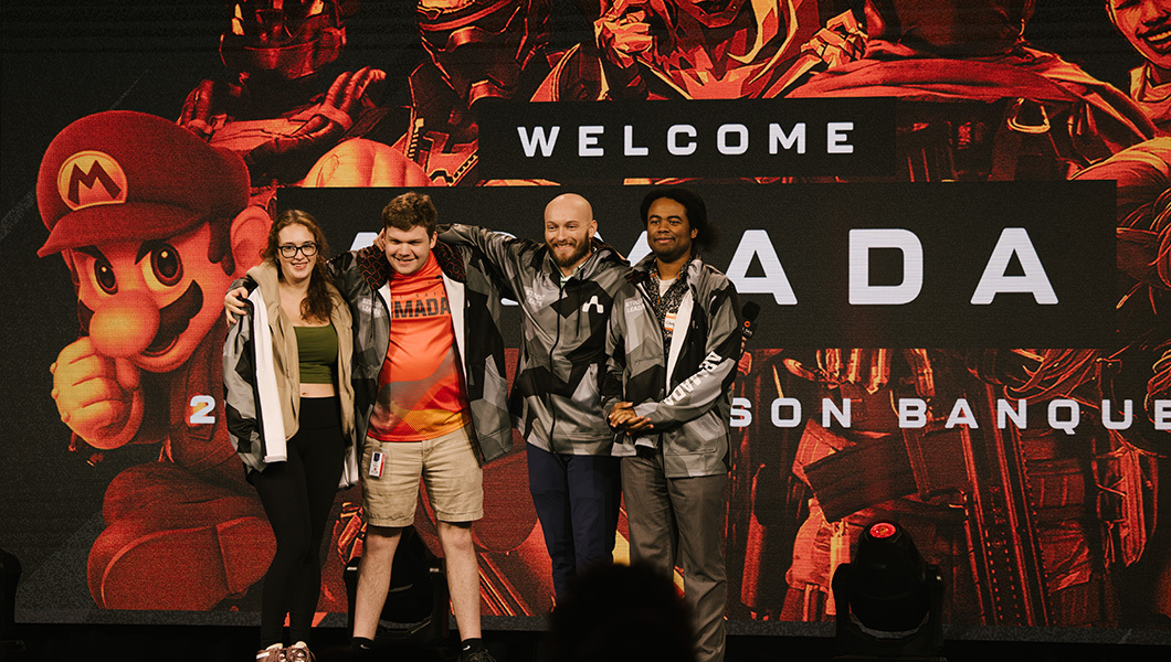 A group of four students stand together arm-in-arm while smiling, they are all wearing gray Armada hooded sweatshirts.