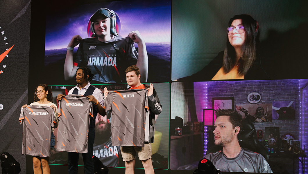 Three students stand on stage while holding up their new jerseys while two online students appear on video on the large screen behind them.