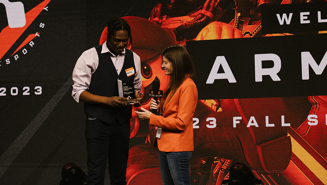 Two people are smiling and standing on stage in the Fortress while one hands an award to the other.