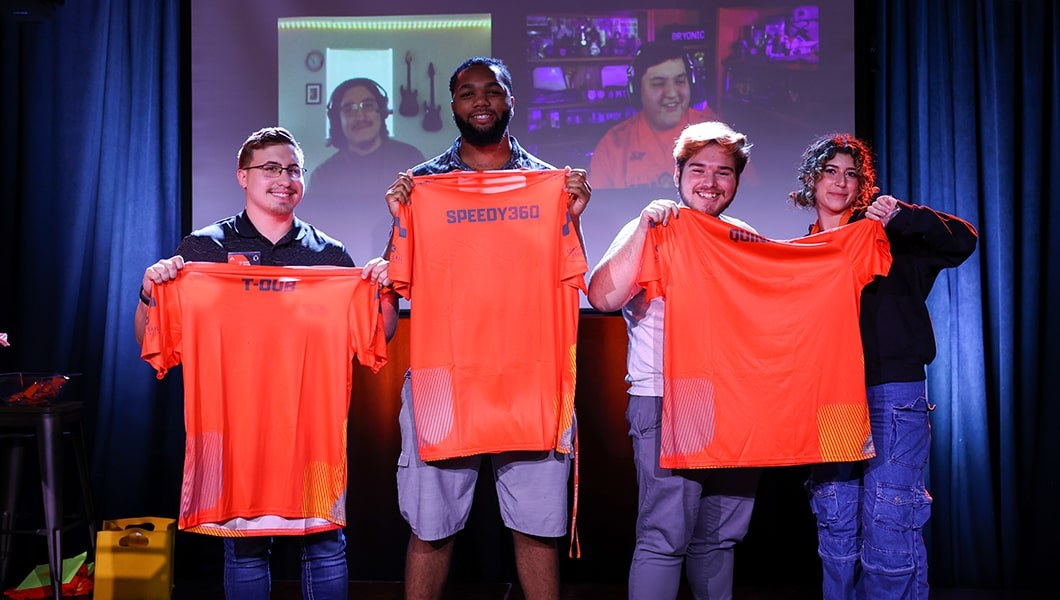 Three Full Sail Armada athletes standing on a stage holding up their new orange Armada jerseys, with two online student appearing on the screen behind them in a video chat.