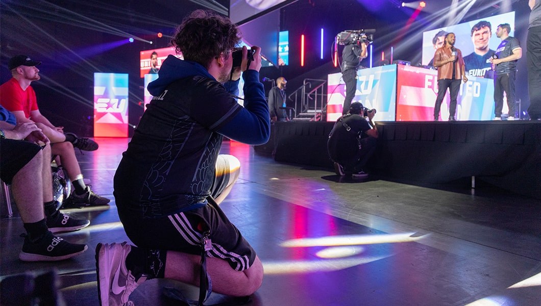 An intern crouches with a camera and takes photos of a host interviewing a player on the HCS Orlando main stage.