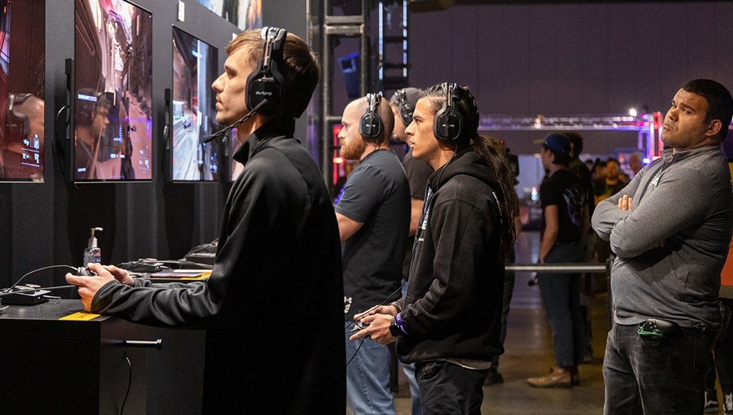 An intern wearing a gaming headset and holding a controller watches gameplay on a screen. Three referees stand beside him.