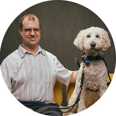 A man wearing a striped button-up shirt and glasses sits on a yellow couch next to his guide dog, a regal, light yellow Goldendoodle.