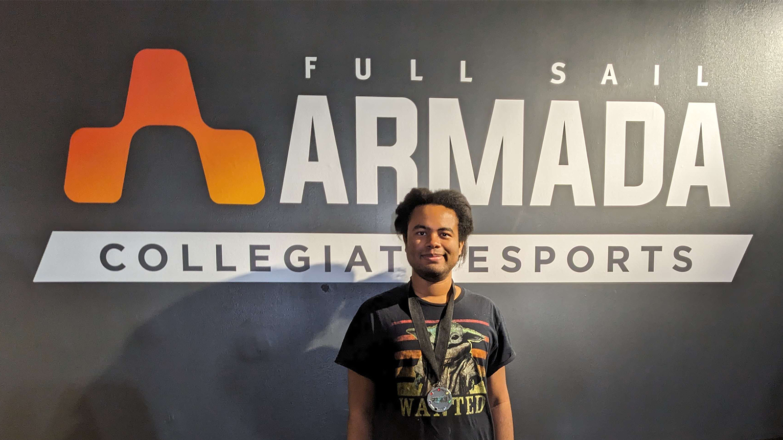 Full Sail Armada athlete Rajah wearing his second place medal while standing in front of a wall mural of the Armada logo.