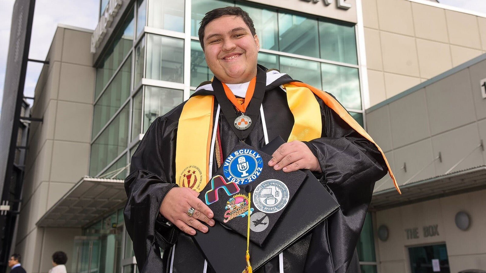 A man with short black hair in a graduation robe with an orange Full Sail sash holding a cap decorated with stickers.