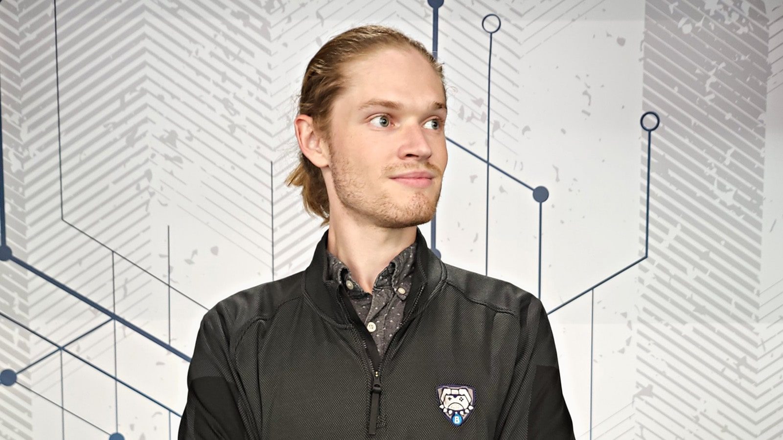 A man with medium-length blonde hair and a black pullover half-zip black jacket featuring a Butler Esports logo on the breast in front of a grey and white geometric backdrop.