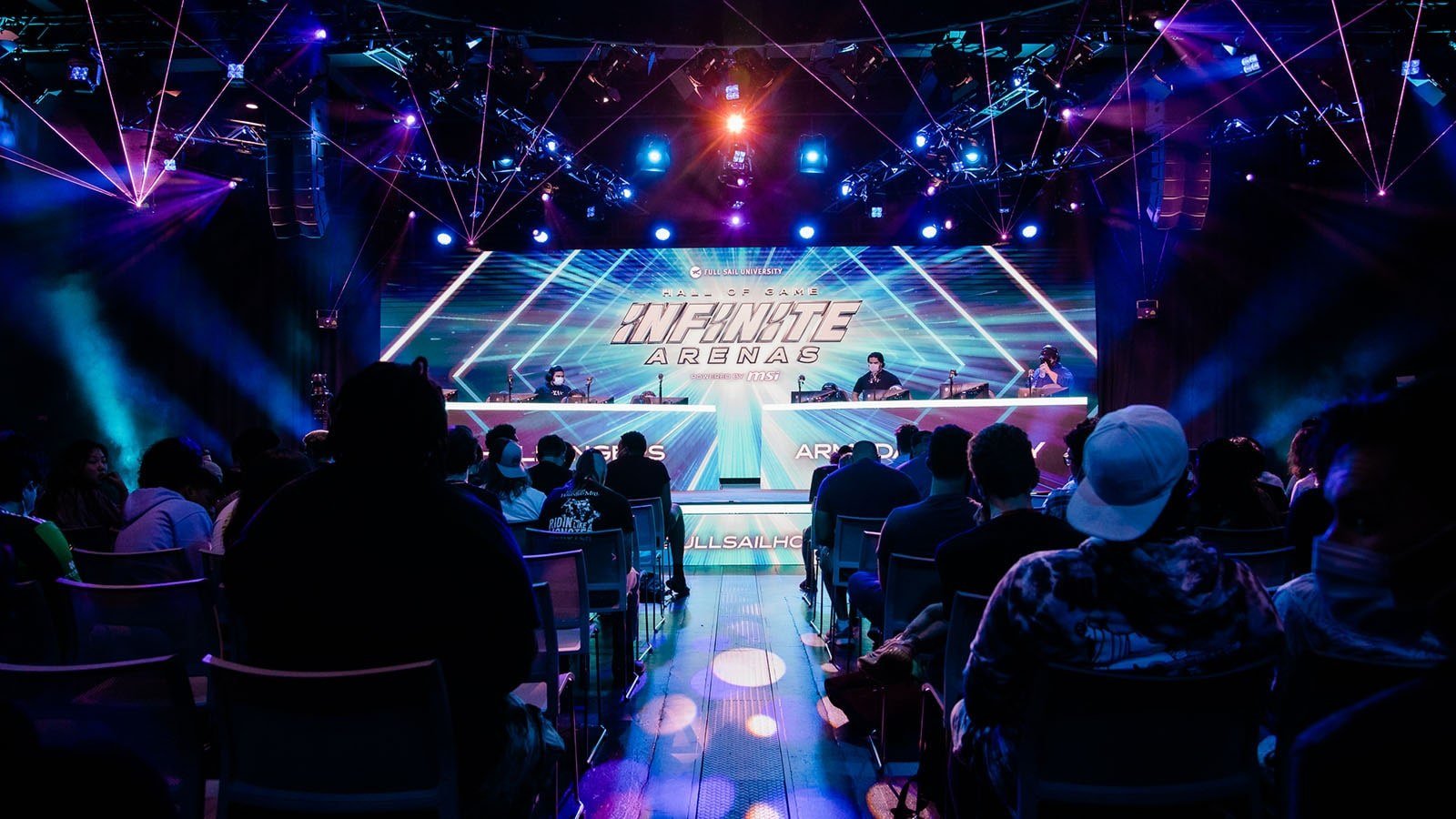 The Full Sail University Orlando Health Fortress’ stage featuring Hall of Game: Infinite Arenas branding in vivid blues, the space is filled with event attendees and illuminated with blue and purple stage lighting.