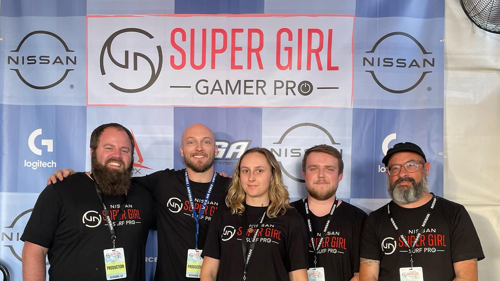 Five people wearing black shirts and badges that read "production" while standing in front of a step and repeat featuring the Super Girl Gamer pro logo and event sponsor logos.