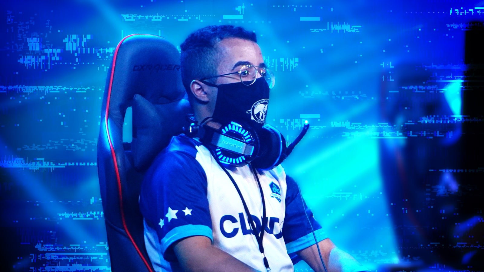 Hector Santos, a young man with short brown hair wearing silver wireframe glasses, a Cloud 9 esports jersey in blue and white, a black face covering, and a gaming headset around his neck, is seated in a gray and orange gaming chair in front of a large blue LED screen on stage at the Fortress.