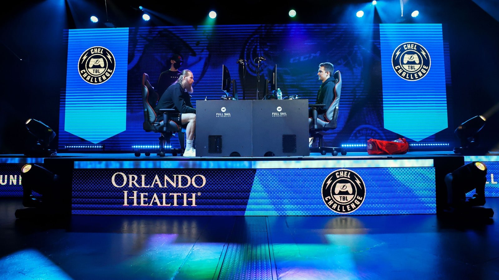 Two players sitting across from one another at gaming stations on a main stage in the Fortress with screens featuring the Orlando Health and Tampa Bay Lightning Bolts Chel Challenge logos against a rich blue background.